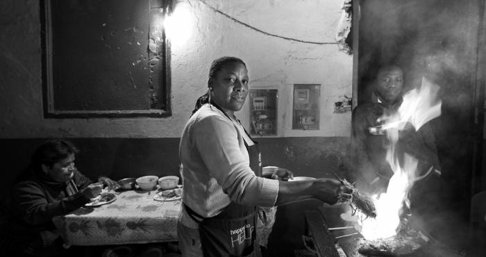 La imagen es un retrato de una mujer afroperuana que mira a la cámara mientras cocina algo a la parrilla.